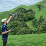 image of man holding a survey drone in lush green field with hill